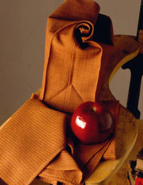 A red apple sits on a beautifully folded piece of fabric from the "Best of Handwoven Yarn Series: A Dozen Projects in 10/2 Pearl Cotton (Printed Version)" by Long Thread Media, draped over a wooden chair. The fabric, reminiscent of a fine tablecloth, features a subtle geometric pattern that adds texture to the arrangement. The background is plain, keeping the focus on the apple and the exquisite fabric.