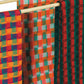 Displayed on a wooden rod are three vibrant woven textiles from the "Best of Handwoven Yarn Series: A Dozen Projects in 10/2 Pearl Cotton" by Long Thread Media. The textile on the left showcases an orange, yellow, blue, and purple checkered pattern. The middle features a design reminiscent of tablecloth towels with orange, white, green, and red checks. The right textile boasts striking green and red stripes accented with hints of black.