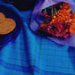 A bouquet of orange and purple flowers wrapped in purple paper is placed on a blue tablecloth. To the left, a copy of "Best of Handwoven Yarn Series: A Dozen Projects in 8/2 Cotton (Printed Version)" by Long Thread Media lies next to three round cookies on a dark blue plate, with a matching dark blue mug partially visible in the background. Napkins patterned in harmonious hues add to the cozy scene.