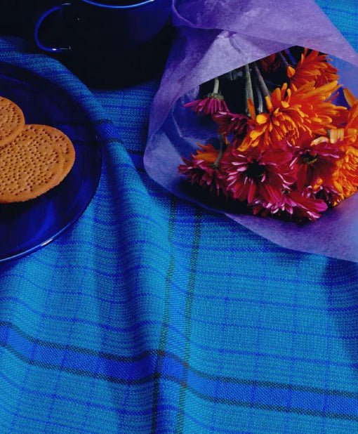 A bouquet of orange and purple flowers wrapped in purple paper is placed on a blue tablecloth. To the left, a copy of "Best of Handwoven Yarn Series: A Dozen Projects in 8/2 Cotton (Printed Version)" by Long Thread Media lies next to three round cookies on a dark blue plate, with a matching dark blue mug partially visible in the background. Napkins patterned in harmonious hues add to the cozy scene.