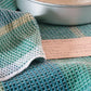 A close-up image of a metallic round baking pan placed on a textured, green and blue checkered cloth that resembles patterned napkins. A handwritten recipe card with visible but illegible text is partially tucked under the cloth. The scene appears to be set for baking alongside Long Thread Media's "Best of Handwoven Yarn Series: A Dozen Projects in 8/2 Cotton (Printed Version).