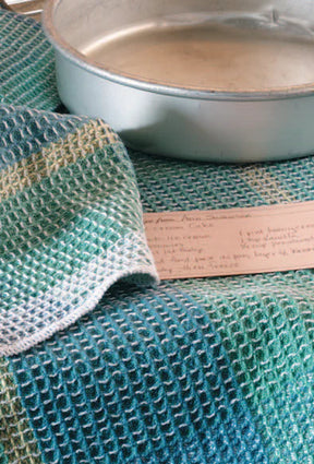 A close-up image of a metallic round baking pan placed on a textured, green and blue checkered cloth that resembles patterned napkins. A handwritten recipe card with visible but illegible text is partially tucked under the cloth. The scene appears to be set for baking alongside Long Thread Media's "Best of Handwoven Yarn Series: A Dozen Projects in 8/2 Cotton (Printed Version).