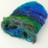 A rolled-up bundle of Demi-Carded Silk Bundles from Indian Silk Shop featuring a mix of vibrant blue, green, and gray hues. The slightly frizzy texture suggests it is hand-dyed or made from multiple fiber types. This exquisite art yarn is perfect for felting projects. The background is plain and white.