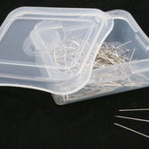 A transparent plastic box, partially open, contains numerous Pleating Pins (350) - Silk Pins without head by Notions. Three of these delicate nickel-plated steel pins lie on the black surface beside the box.