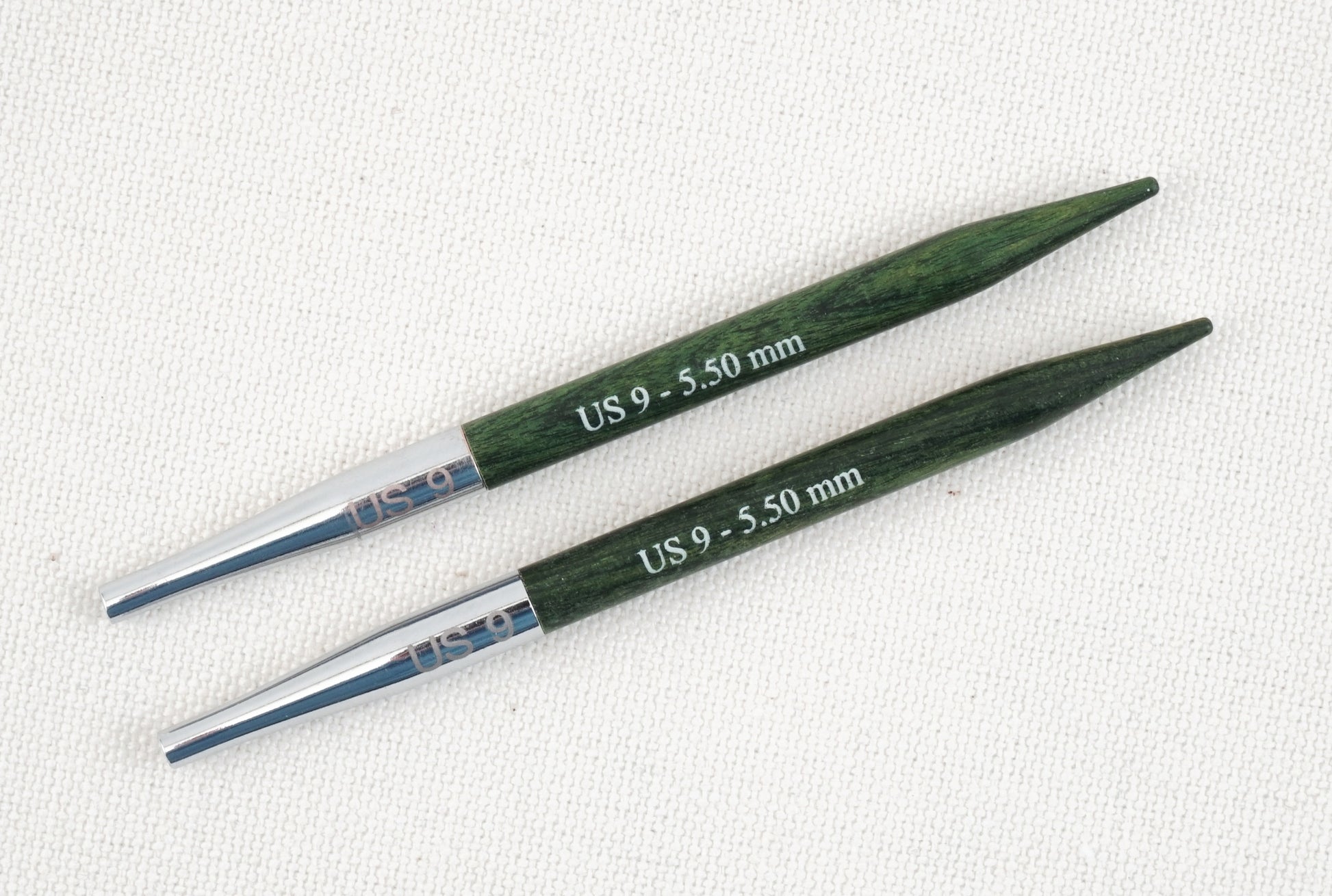 Two Dreamz Interchangeable Knitting Needle Tips by Accessories Unlimited, in dark green wood and inscribed with "US 9 - 5.50 mm" on the tips and "US 9" on the metal connector end, are arranged on a beige fabric background—perfect for knitting hats with smooth joins.