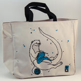 The Otter Project Tote by Mum n Sun Ink from Bonnie Bishoff is a beige Envirotote canvas bag with black handles, featuring a whimsical illustration of an otter knitting a blue scarf surrounded by blue bubbles. A small penguin logo in the bottom right corner adds extra charm. Perfect for carrying your knitting essentials or shawl pins.