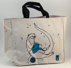 The Otter Project Tote by Mum n Sun Ink from Bonnie Bishoff is a beige Envirotote canvas bag with black handles, featuring a whimsical illustration of an otter knitting a blue scarf surrounded by blue bubbles. A small penguin logo in the bottom right corner adds extra charm. Perfect for carrying your knitting essentials or shawl pins.