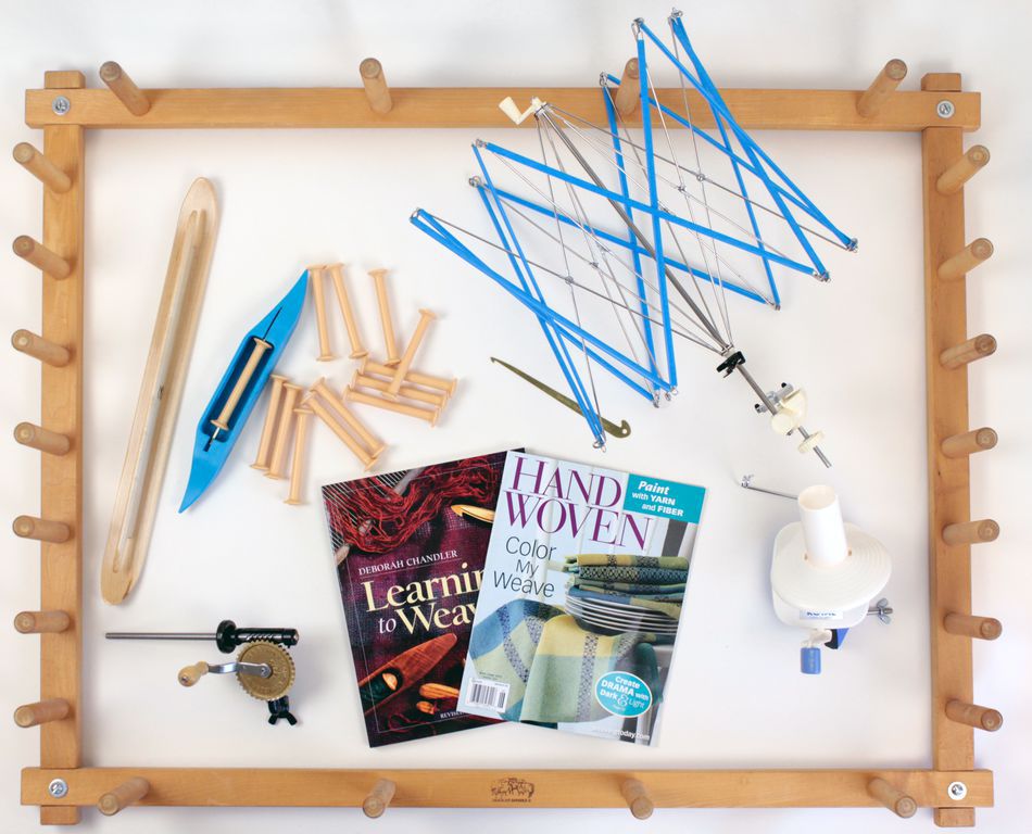 A wooden frame setup for weaving is laid out on a white surface. Various weaving tools including shuttles, spools, and an umbrella swift are inside the frame. Nearby magazines titled "Learn to Weave" and "Handwoven" are also present alongside a Halcyon Yarn Quick Start Floor Loom Accessory Package.