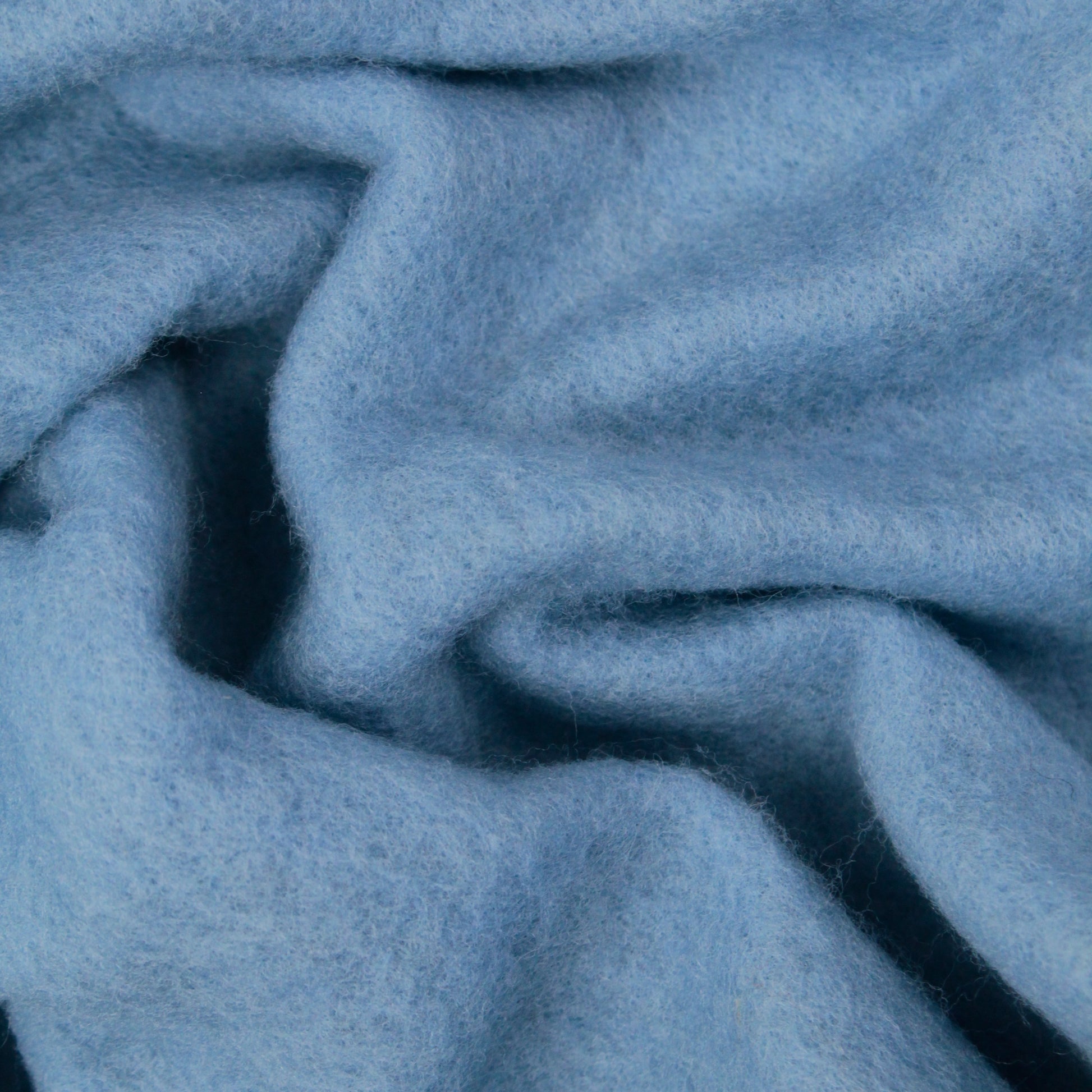 A close-up image showcases the soft, light blue fabric of Europa Wools Ltd's Merino Prefelt, featuring a slightly fuzzy texture. Gentle wrinkles create subtle folds and shadows across the cozy and warm material, making it an ideal choice for nuno felting projects.