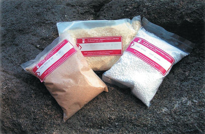 Three plastic bags filled with different substances are displayed against a rocky background. The left bag contains brown powder, the middle bag holds beige shavings, and the right bag has white granules. Each bag bears a red and white label with text for G & K Craft's Pro Chem Flakes, ideal for curing or reduction prevention.