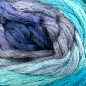 A close-up of soft, thick strands from the Cumulus Cotton by Juniper Moon Farm, available through Knitting Fever / Euro Yarns, intertwined in shades of light blue, dark blue, teal, and gray with durable chainette construction. The texture appears fluffy and smooth, suggesting a cozy and warm material ideal for knitting weightless accessories or crocheting.