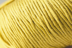 A close-up photograph of a yellow summer yarn ball. The tightly wound strands showcase the texture and detail of the Jo Sharp Soho Summer DK Cotton fibers from Kingfisher Yarn & Fibre. The close-up perspective highlights the fine and consistent weave of this lightweight cotton yarn.