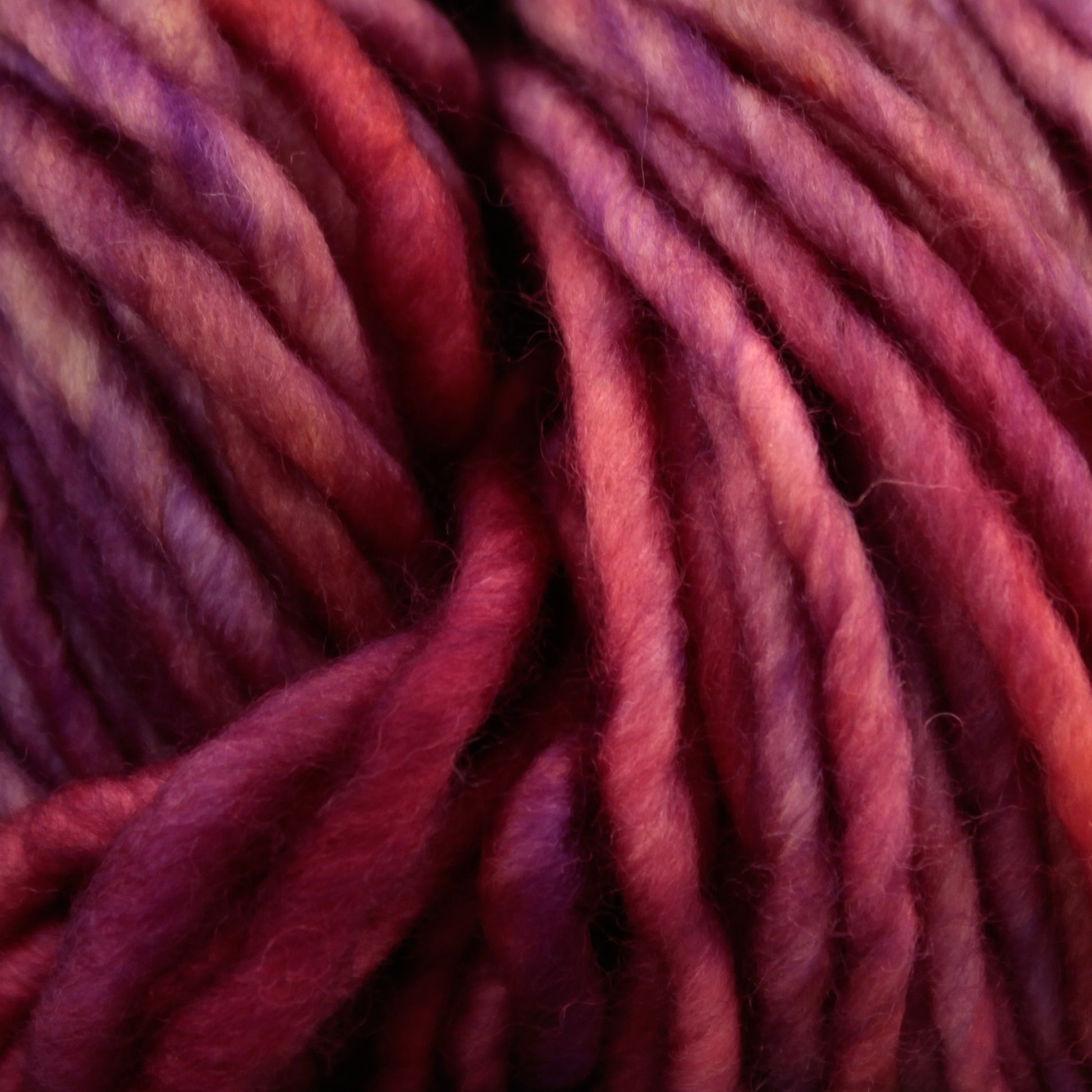 Close-up image of thick, multi-colored Malabrigo Mecha yarn in shades of red, purple, and pink. The kettle-dyed fibers from Malabrigo Yarn are twisted and slightly fuzzy, forming a textured, intertwined pattern.