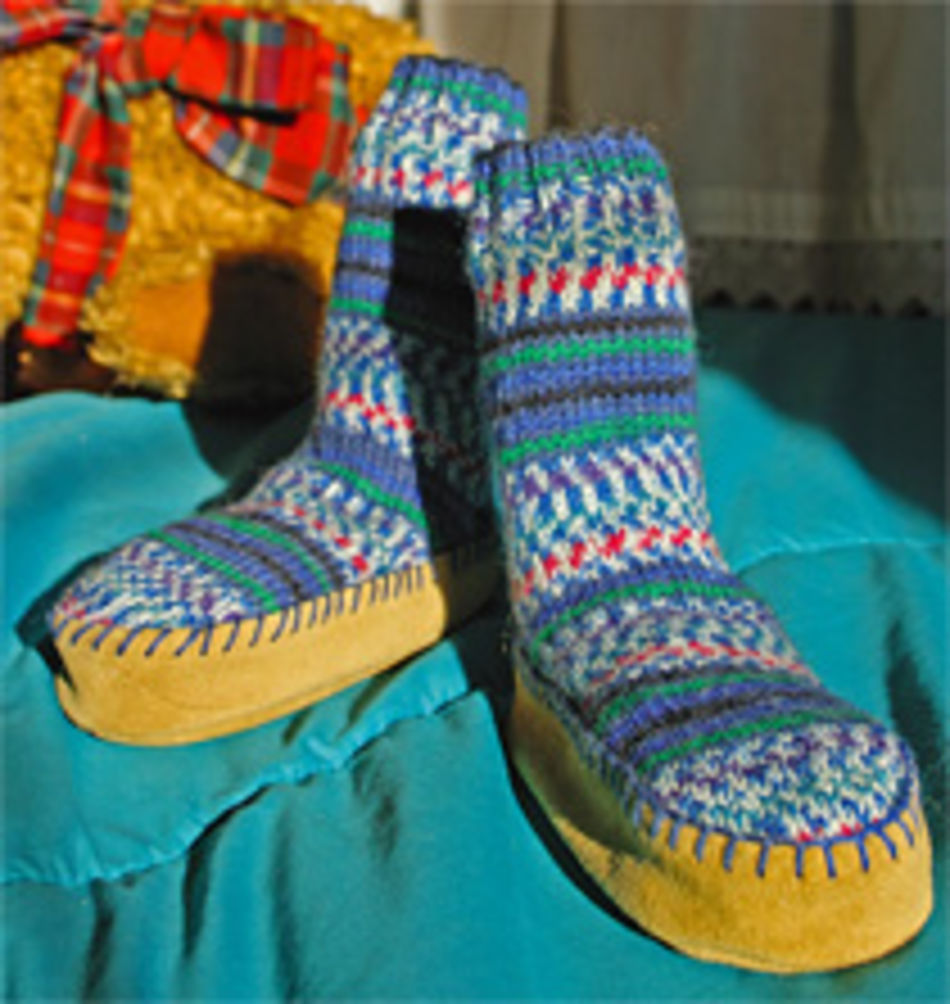 A pair of colorful, patterned knitted slippers with soft soles sit on a teal blanket. The slippers, featuring various hues of blue, green, and white, have Bryson Distributing Inc.'s Suede Leather Slipper Soles made of genuine suede leather. In the background is a decorative plaid item.