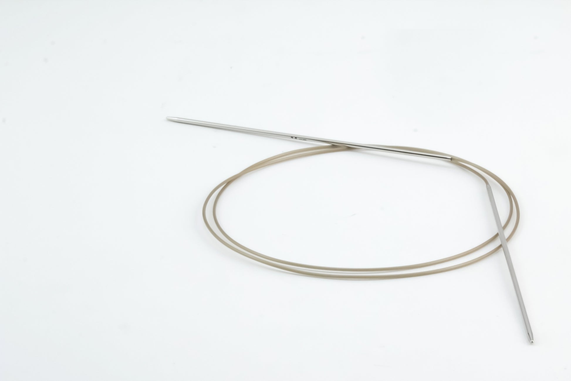 A pair of Skacel Addi Turbo Circular Knitting Needles, featuring coiled, beige tubing with metal tips, is arranged in an elongated loop on a pristine white background. One tip is slightly elevated while the other rests flat on the surface.