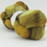 A skein of soft, hand-dyed merino wool in a mix of earthy tones including green, yellow, brown, and hints of orange. The label reads "Nube by Malabrigo". The fiber has a fluffy, smooth texture, suggesting it's suitable for spinning or felting projects.