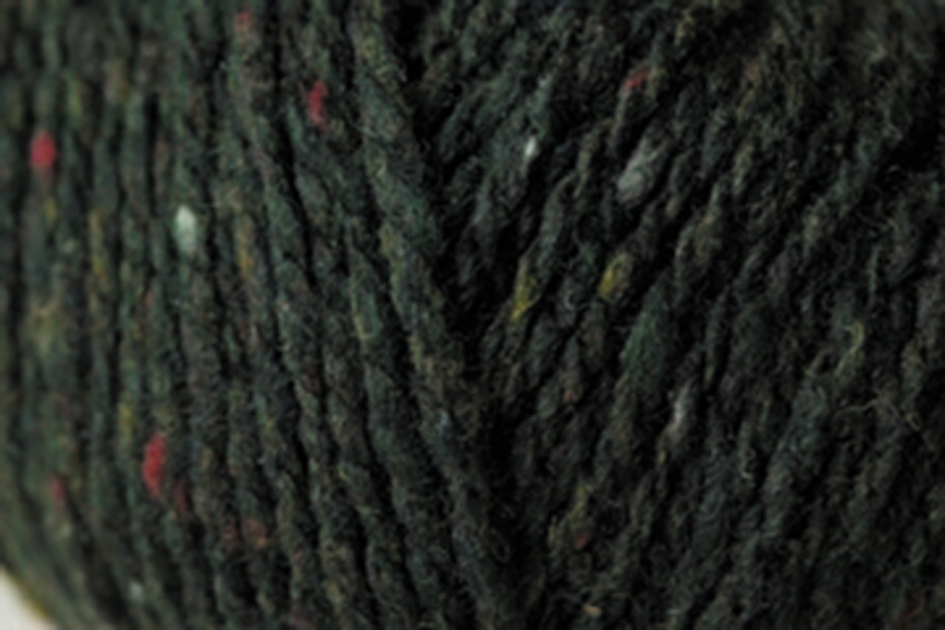 Close-up image of dark green Jo Sharp Silkroad DK Tweed yarn by Kingfisher Yarn & Fibre, featuring small flecks of red, white, and yellow. The texture of the yarn appears slightly coarse and thick, making it suitable for knitting or crocheting.