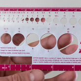 A hand holds the Accessories Unlimited Knit-Chek gauge tool, a handy device that includes knitting needle size check guides, a ruler in inches and centimeters, and openings to measure stitch size. This rectangular tool features various circular cutouts and printed measurement markings for accurate crafting.