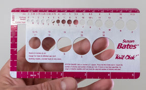 A hand holds the Accessories Unlimited Knit-Chek gauge tool, a handy device that includes knitting needle size check guides, a ruler in inches and centimeters, and openings to measure stitch size. This rectangular tool features various circular cutouts and printed measurement markings for accurate crafting.