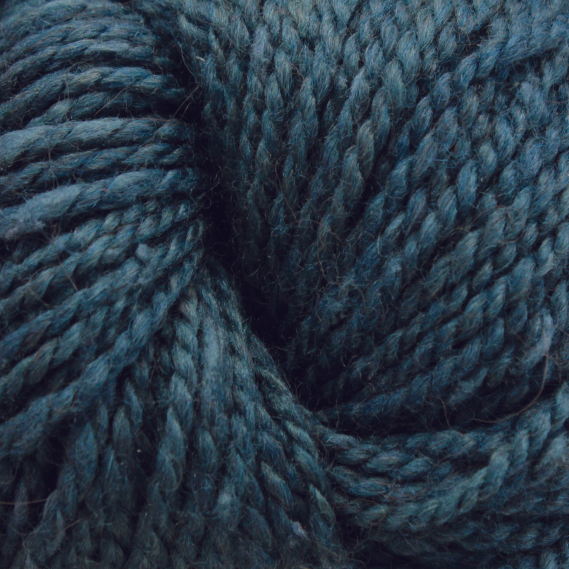 Close-up of dark teal Acadia by The Fiber Company yarn, tightly twisted. The fibers appear soft and textured, with a subtle tweed effect adding depth to the varying shades of teal. The thick and robust yarn evokes natural scenic beauty, making it perfect for knitting or crocheting projects by The Fibre Co. (UK) Ltd.