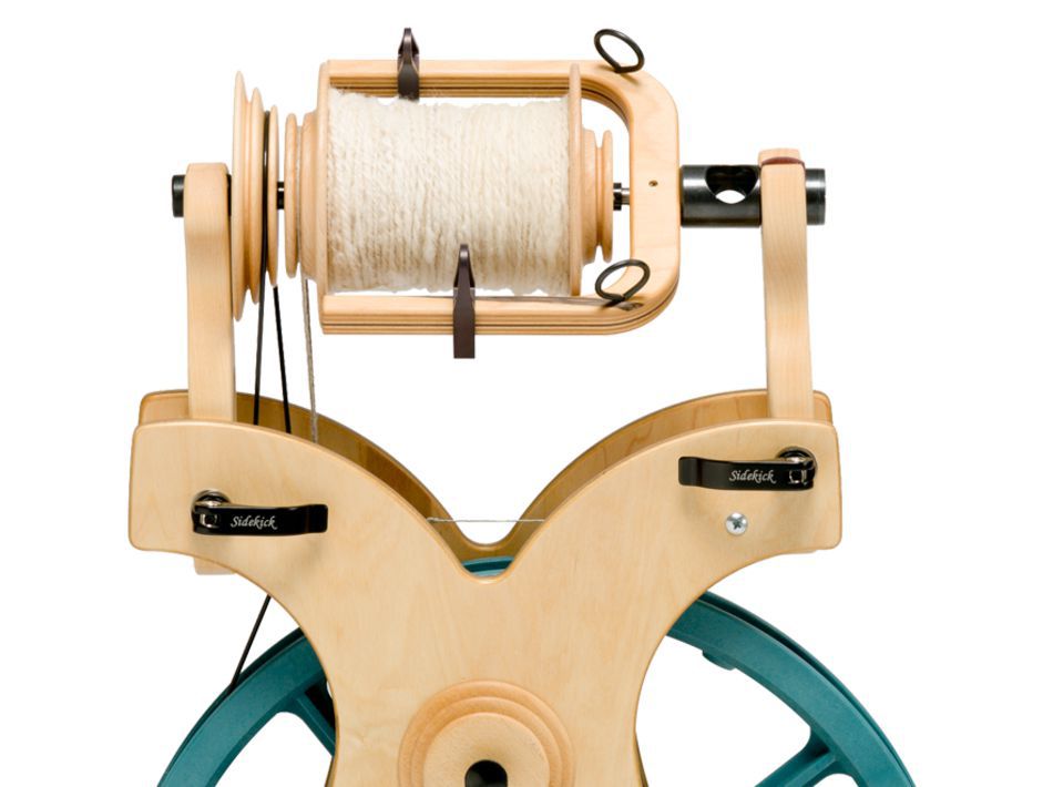 A close-up view of the Schacht Bulky Plyer Flyer Package featuring wooden parts and a spool of white yarn. The wheel has metal hooks and tension knobs, with a segment of a blue wheel visible beneath the wooden structure. The brand "Schacht Spindle Co." is visible on the wheel.