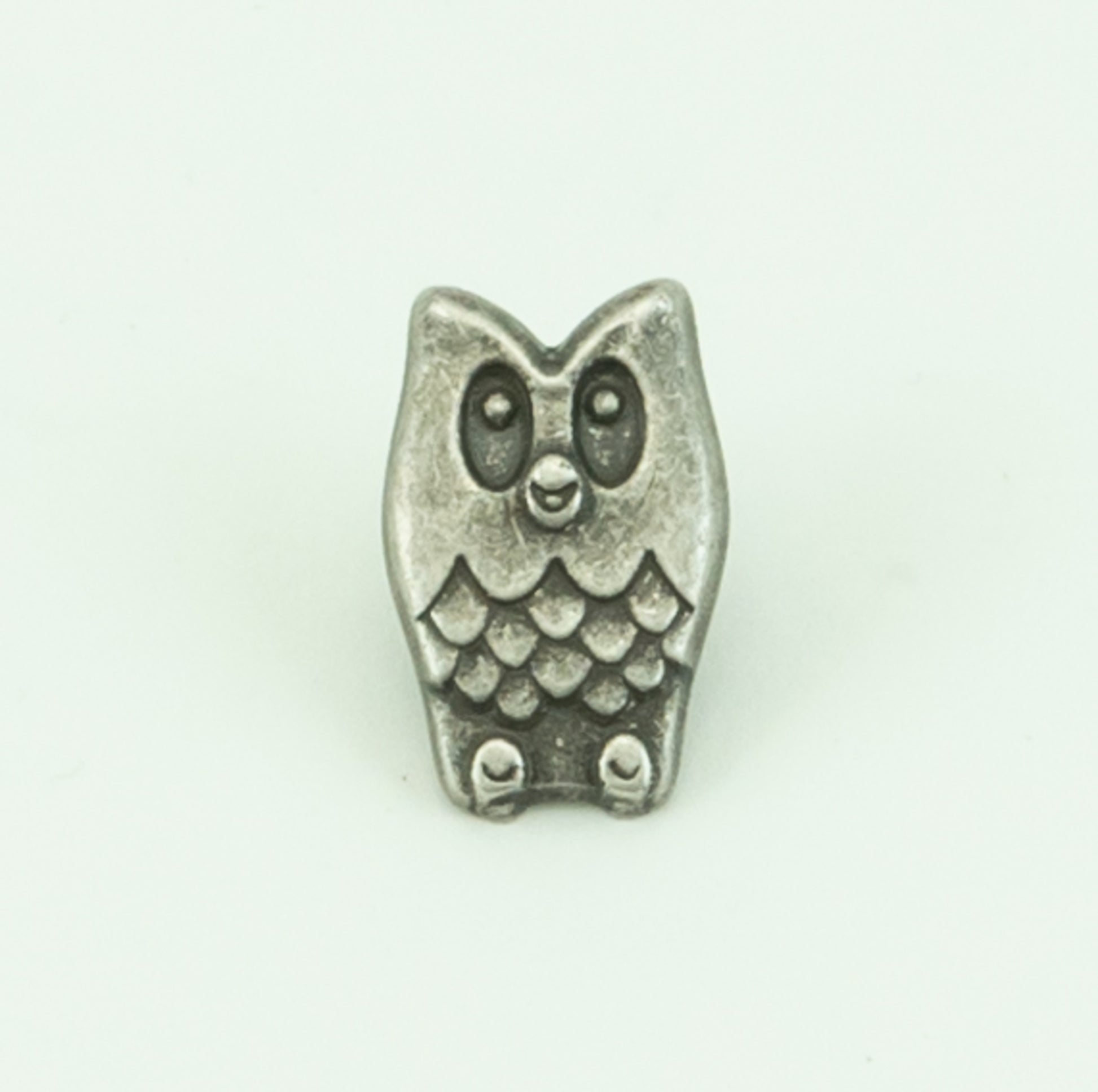 The Pewterette Button by Buttons Etc. is a small, silver charm shaped like an owl, featuring big round eyes and an intricately detailed feather pattern reminiscent of a metal button. Measuring 11/16" in diameter, this charm has a slightly tarnished look and is set against a plain white background.