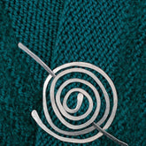 A close-up image of Carolyn Jackson's Aluminum Shawl Pin pinned on teal knitted fabric. The shawl pin has a simple, elegant design with concentric circles and a pin running through the center. The textured fabric provides a contrasting background.