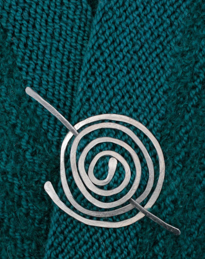 A close-up image of Carolyn Jackson's Aluminum Shawl Pin pinned on teal knitted fabric. The shawl pin has a simple, elegant design with concentric circles and a pin running through the center. The textured fabric provides a contrasting background.