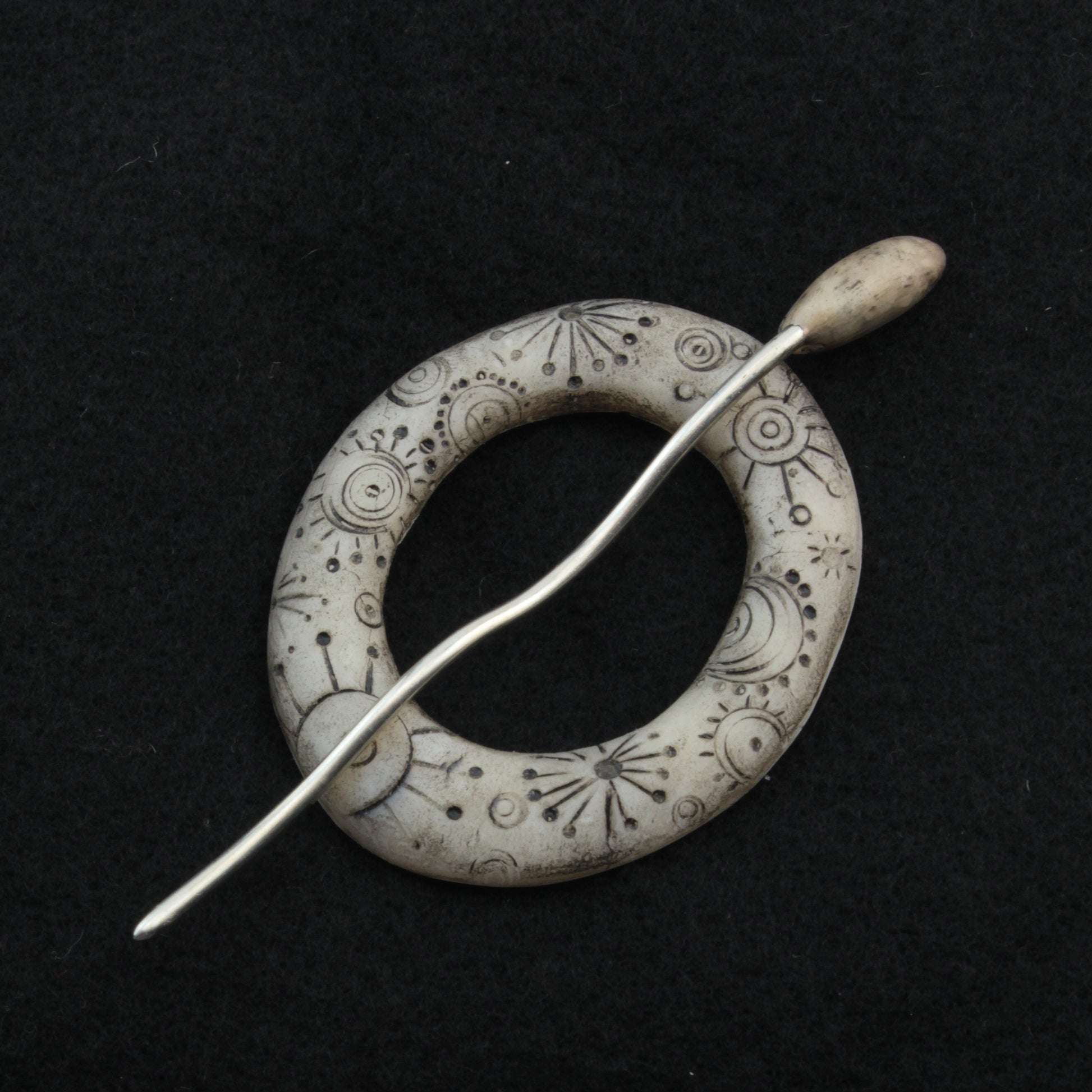 The Carved Ring Shawl Pin by Bonnie Bishoff Designs is a handcrafted creation featuring a circular, stone-like base adorned with intricate engraved patterns, including stars and abstract designs. A slender, slightly curved silver pin runs through the center, set against a dark fabric background. This piece from Bonnie Bishoff is perfect for securing lightweight shawls or adding a decorative touch to knit wearables.