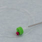 The Accessories Unlimited Clover Circular Needle & Stitch Holder features a clear plastic ring encircling a green and red bead, connected to a metallic rod with a sharp tip, resembling an intricate stitch holder, resting on a light gray surface.