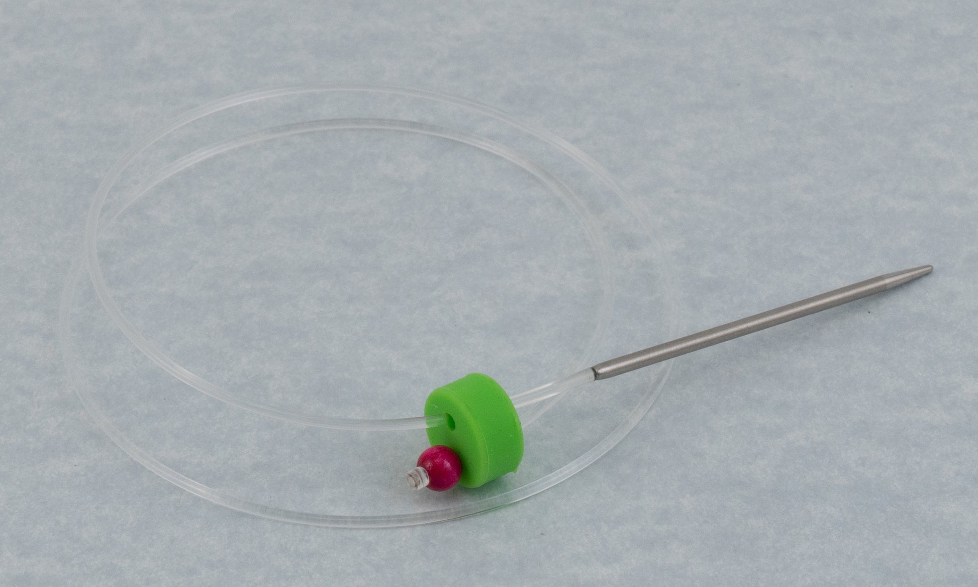 The Accessories Unlimited Clover Circular Needle & Stitch Holder features a clear plastic ring encircling a green and red bead, connected to a metallic rod with a sharp tip, resembling an intricate stitch holder, resting on a light gray surface.