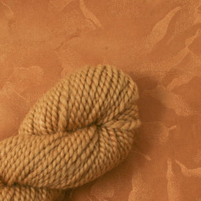 A close-up of a skein made with Earthues Cutch Extract thick, light brown yarn resting on a matching light brown textured surface. Hints of reds and yellows subtly weave through the tightly twisted, plump, and soft strands. The background showcases a subtle, abstract marbled pattern that complements the color of this Earthues yarn.