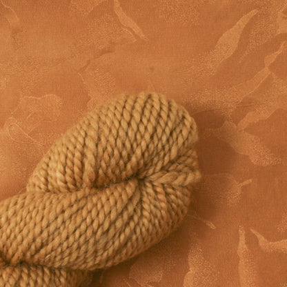 A close-up of a skein made with Earthues Cutch Extract thick, light brown yarn resting on a matching light brown textured surface. Hints of reds and yellows subtly weave through the tightly twisted, plump, and soft strands. The background showcases a subtle, abstract marbled pattern that complements the color of this Earthues yarn.