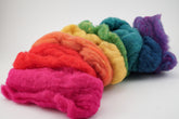 A collection of Halcyon Yarn's Harrisville Fiber Batts arranged in a gradient from pink to red, orange, yellow, green, blue, and purple creates a vibrant rainbow effect. Perfect for felting projects or creating multi-color bags, the wool fibers appear soft and fluffy against a plain white background.