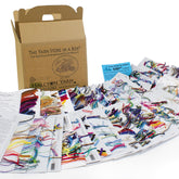 A variety of colorful yarn samples are displayed on multiple pages of a booklet. A cardboard box labeled "Yarn Store in a Box® - the classic design collection" from Halcyon Yarn is in the background. Additionally, small instructional booklets and a promotional card for fiber artists are included in the scene.