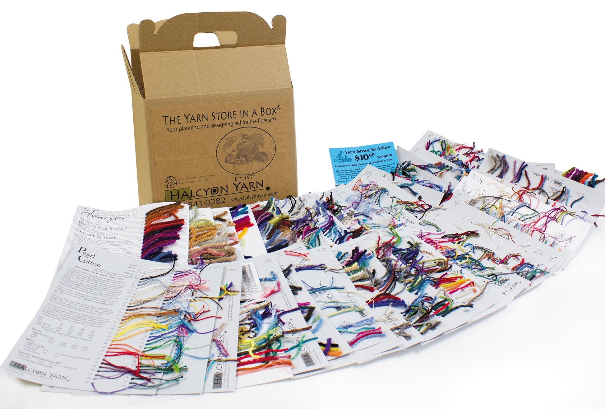 A variety of colorful yarn samples are displayed on multiple pages of a booklet. A cardboard box labeled "Yarn Store in a Box® - the classic design collection" from Halcyon Yarn is in the background. Additionally, small instructional booklets and a promotional card for fiber artists are included in the scene.