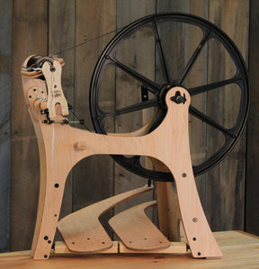 The Flatiron Spinning Wheel, Double-Treadle by Schacht from Schacht Spindle Co. stands against a rustic wooden backdrop, showcasing its sturdy frame and large black adjustable drive wheel. This exquisite wheel includes pedals and various mechanisms for spinning fiber into yarn, while the natural wood finish accentuates the craftsmanship of this highly customizable spinning wheel.