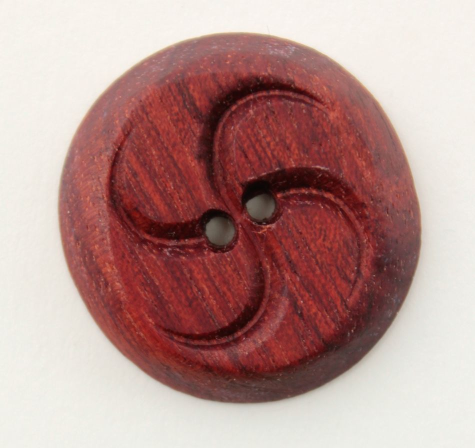 A close-up photo of the Wood Button by Alosada, featuring a reddish-brown tone and crafted from all-natural local materials. The button, produced by Alosada LLC, has two holes in the center and a distinctive swirling, propeller-like carved design around the holes. The visible wood grain adds texture to the button's surface.