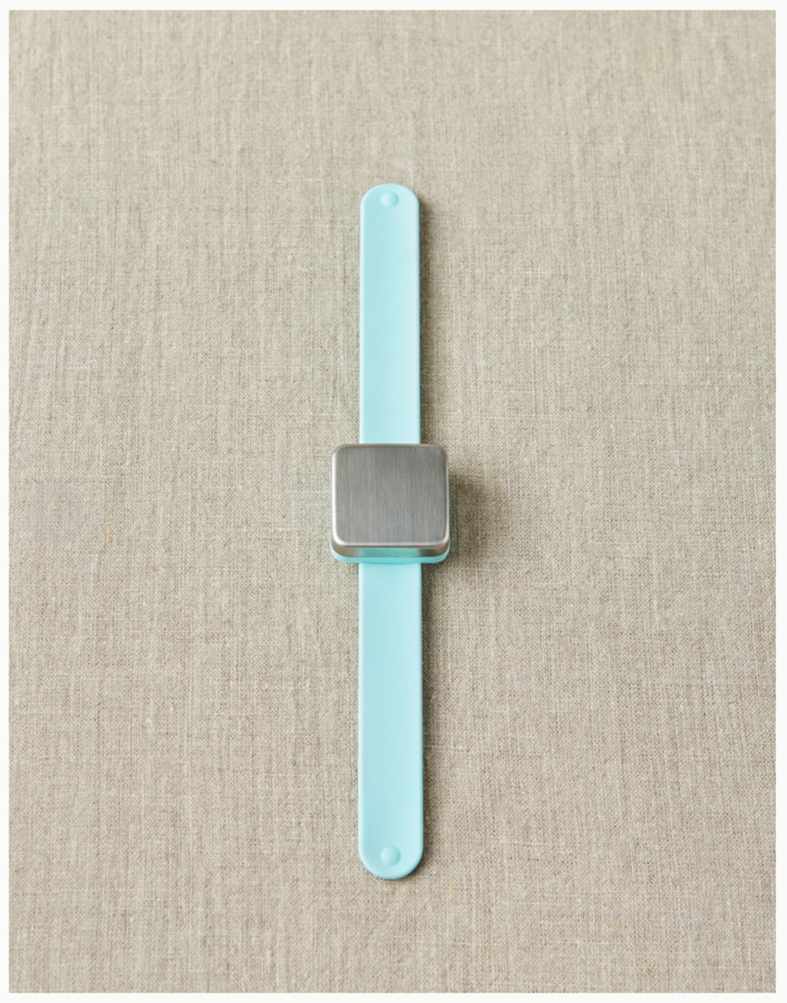 The Maker's Keep Magnetic Bracelet by Cocoknits, featuring a minimalist design with a rectangular silver face and a light blue silicone slap bracelet, is laid flat on a beige fabric background. The rectangular silver face boasts a brushed metal finish with no visible markings or details.