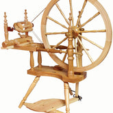 The Kromski Polonaise Spinning Wheel by Kromski North America is a traditional wooden spinning wheel, featuring a large wheel on the right, a smaller wheel on the left, and a foot pedal at the base. Its intricate construction includes multiple spindles and supports, all crafted from light-colored European alder and birch wood.