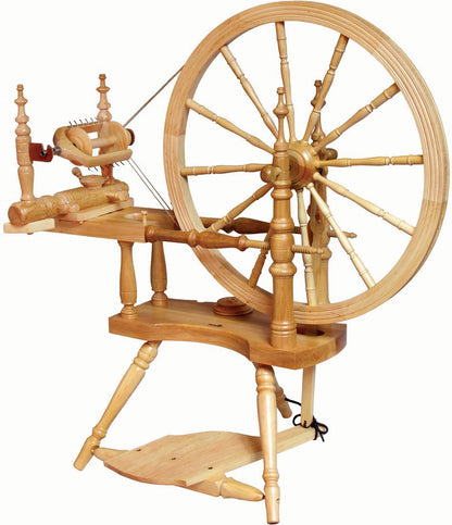 The Kromski Polonaise Spinning Wheel by Kromski North America is a traditional wooden spinning wheel, featuring a large wheel on the right, a smaller wheel on the left, and a foot pedal at the base. Its intricate construction includes multiple spindles and supports, all crafted from light-colored European alder and birch wood.