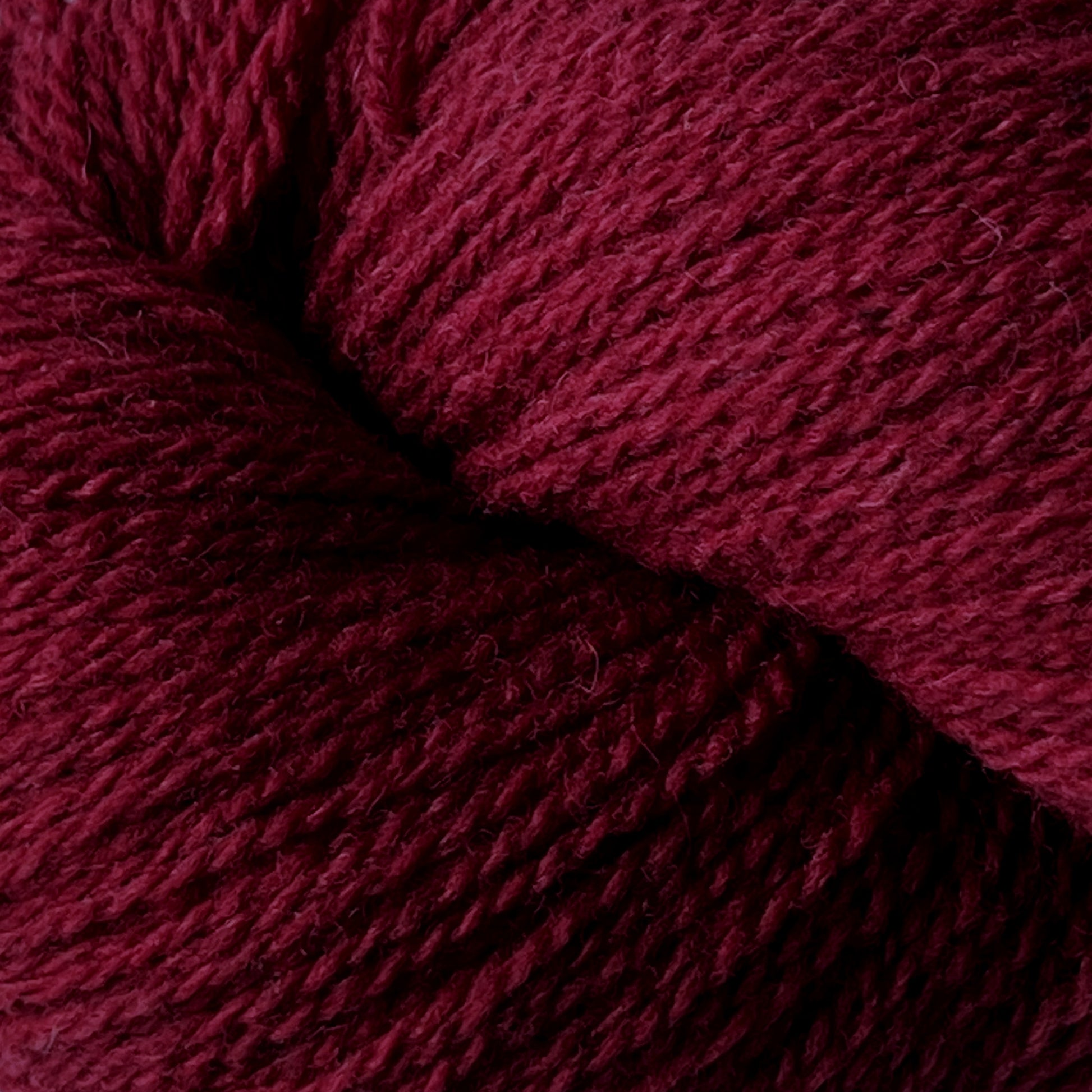 A close-up image of a bundle of dark red yarn. The yarn fibers, made from Patagonia Organic Merino by Juniper Moon Farm, are tightly twisted together, showcasing the texture and thickness of the material. The deep red hue gives this GOTS certified yarn from Knitting Fever / Euro Yarns a rich, warm appearance.