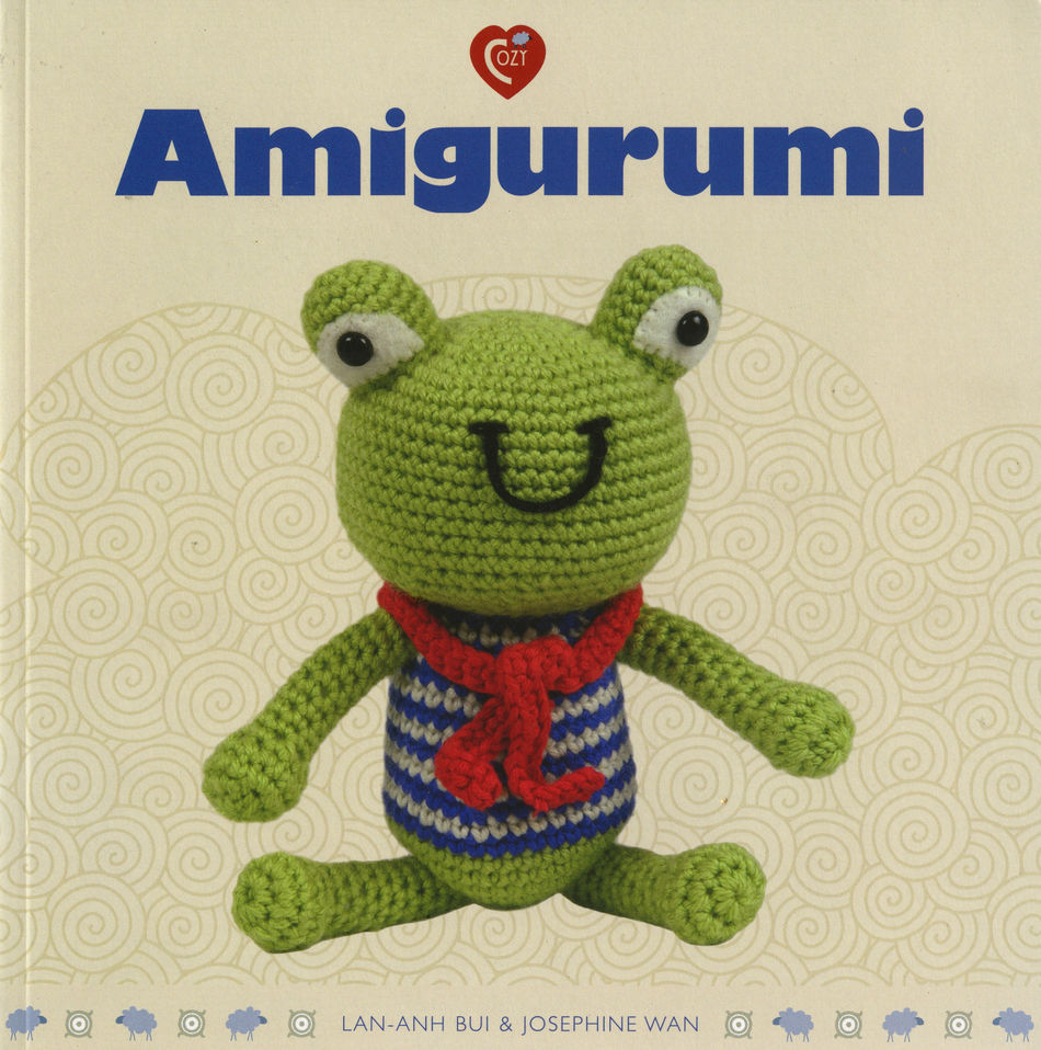 The book cover for "Amigurumi" by Ingram Content features a charming crocheted stuffed toy—a green frog with bulging eyes, wearing a red scarf and a blue and white striped shirt. The background is a light beige color adorned with swirling patterns, and the authors Lan-Anh Bui & Josephine Wan are listed at the bottom.