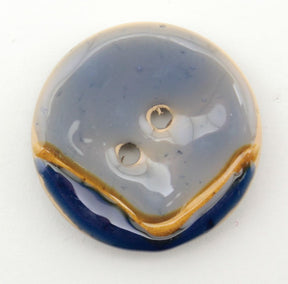 A Coco Boomerang 1 1/8" Button from Buttons Etc. is a round ceramic button with a glazed finish that showcases a marbled design in shades of gray, blue, and gold. It features two central sewing holes and exudes an elegance reminiscent of shellac buttons.