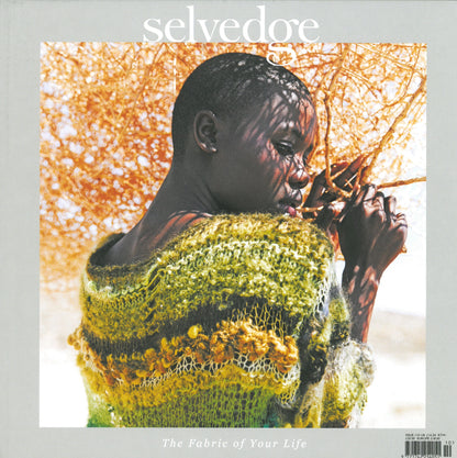 A person wearing a multicolored, textured knit garment leans against a large, dry bush. The text "Selvedge - Issue 110: To Dye For" appears at the top, and "The Fabric of Your Life" is written near the bottom. The light, blurred background emphasizes the mood-lifting Marrs Green in their attire.