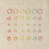 A collection of colorful knitting accessories, including Cocoknits' "Flight of Stitch Markers" made of nylon-coated steel, arranged in a grid pattern on a beige fabric background. The markers come in various shapes such as circles, triangles, and spirals in vibrant colors like red, orange, yellow, green, blue, and purple.