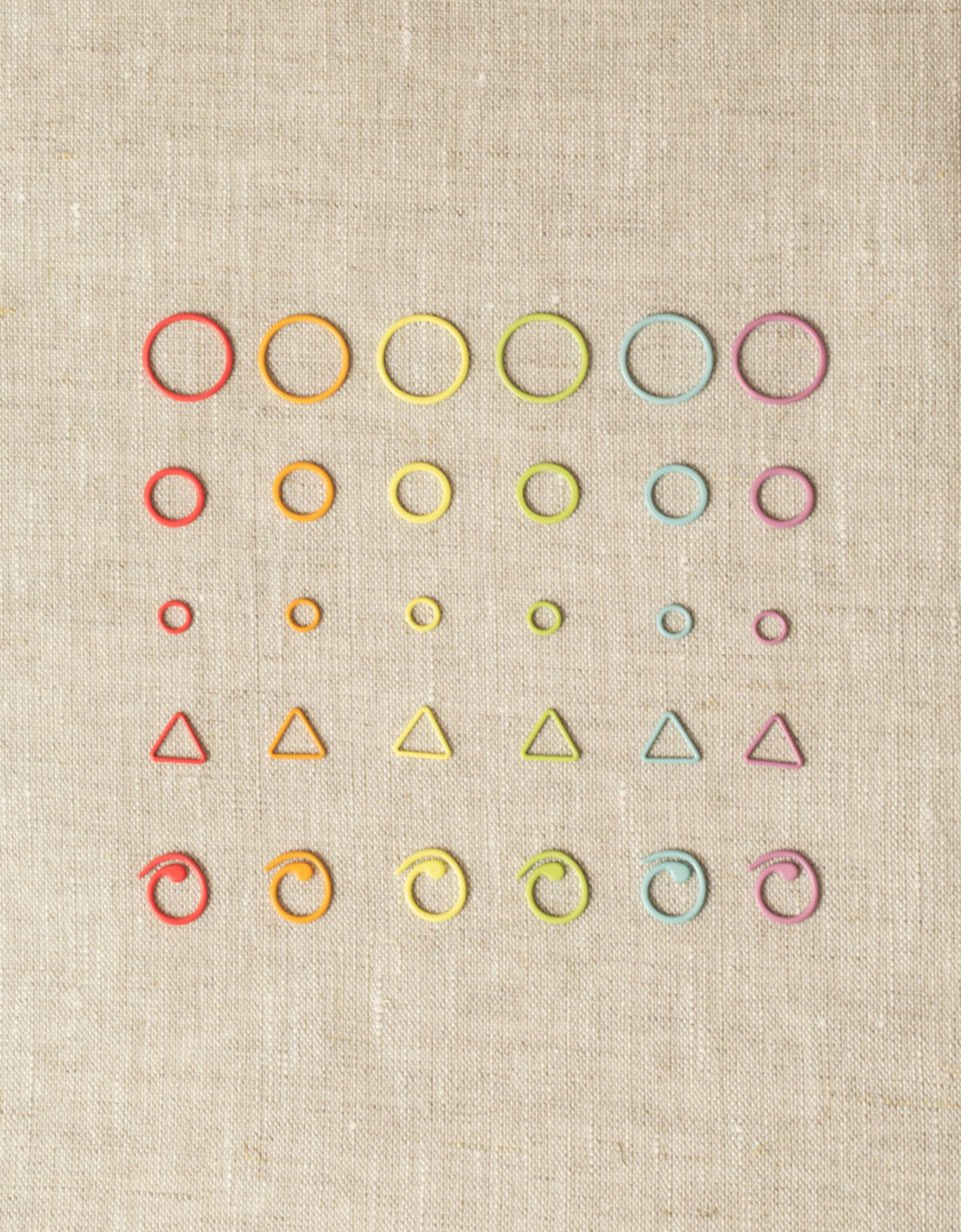 A collection of colorful knitting accessories, including Cocoknits' "Flight of Stitch Markers" made of nylon-coated steel, arranged in a grid pattern on a beige fabric background. The markers come in various shapes such as circles, triangles, and spirals in vibrant colors like red, orange, yellow, green, blue, and purple.