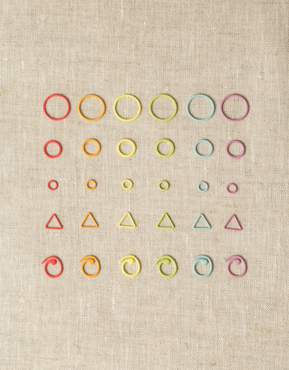 A collection of colorful knitting accessories, including Cocoknits' "Flight of Stitch Markers" made of nylon-coated steel, arranged in a grid pattern on a beige fabric background. The markers come in various shapes such as circles, triangles, and spirals in vibrant colors like red, orange, yellow, green, blue, and purple.