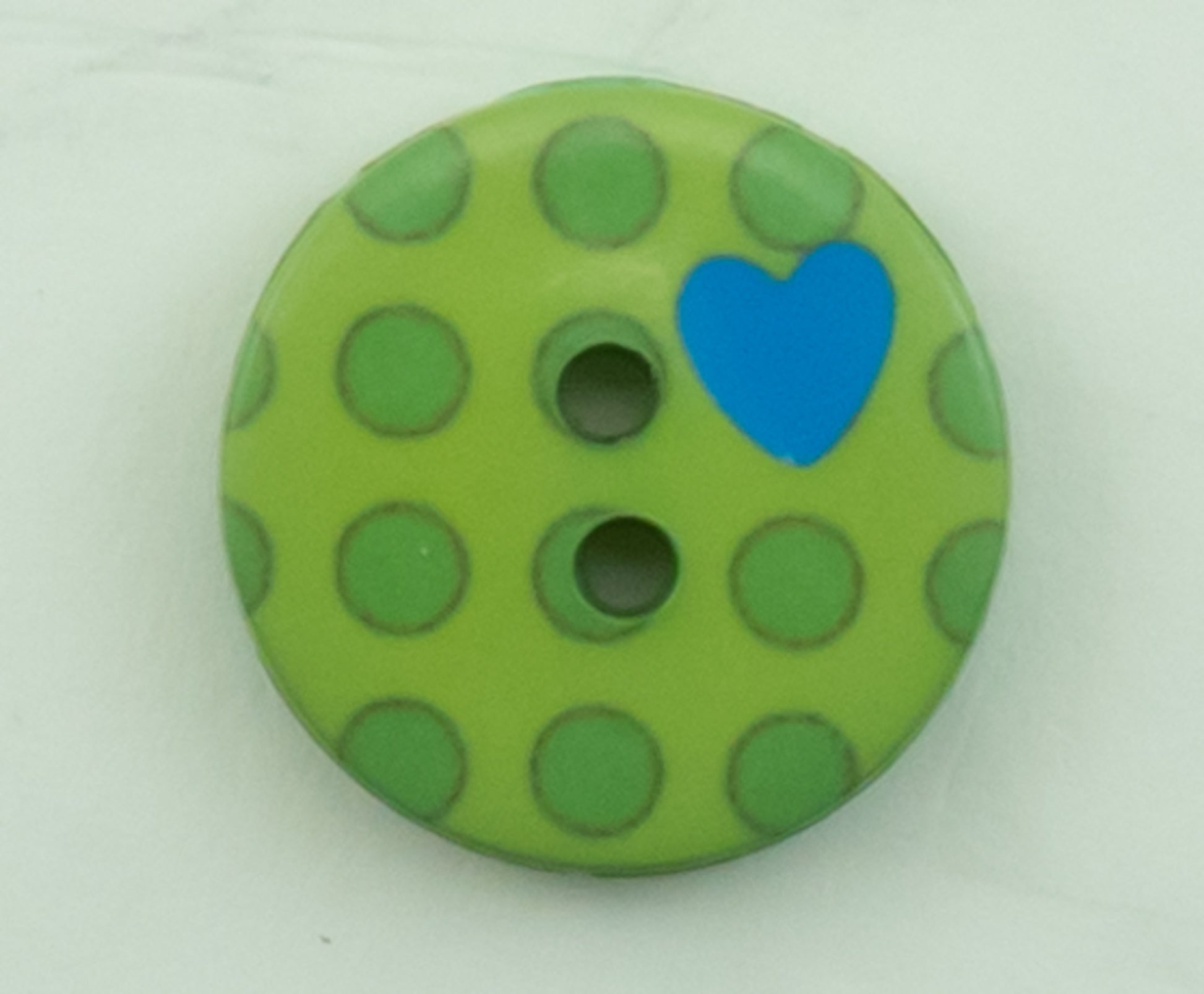 The Dots Button by Buttons Etc. is a two-hole button featuring a green base adorned with dark green polka dots, measuring 5/8" in plastic diameter. A small blue heart is positioned near the top right corner, set against a plain white background.