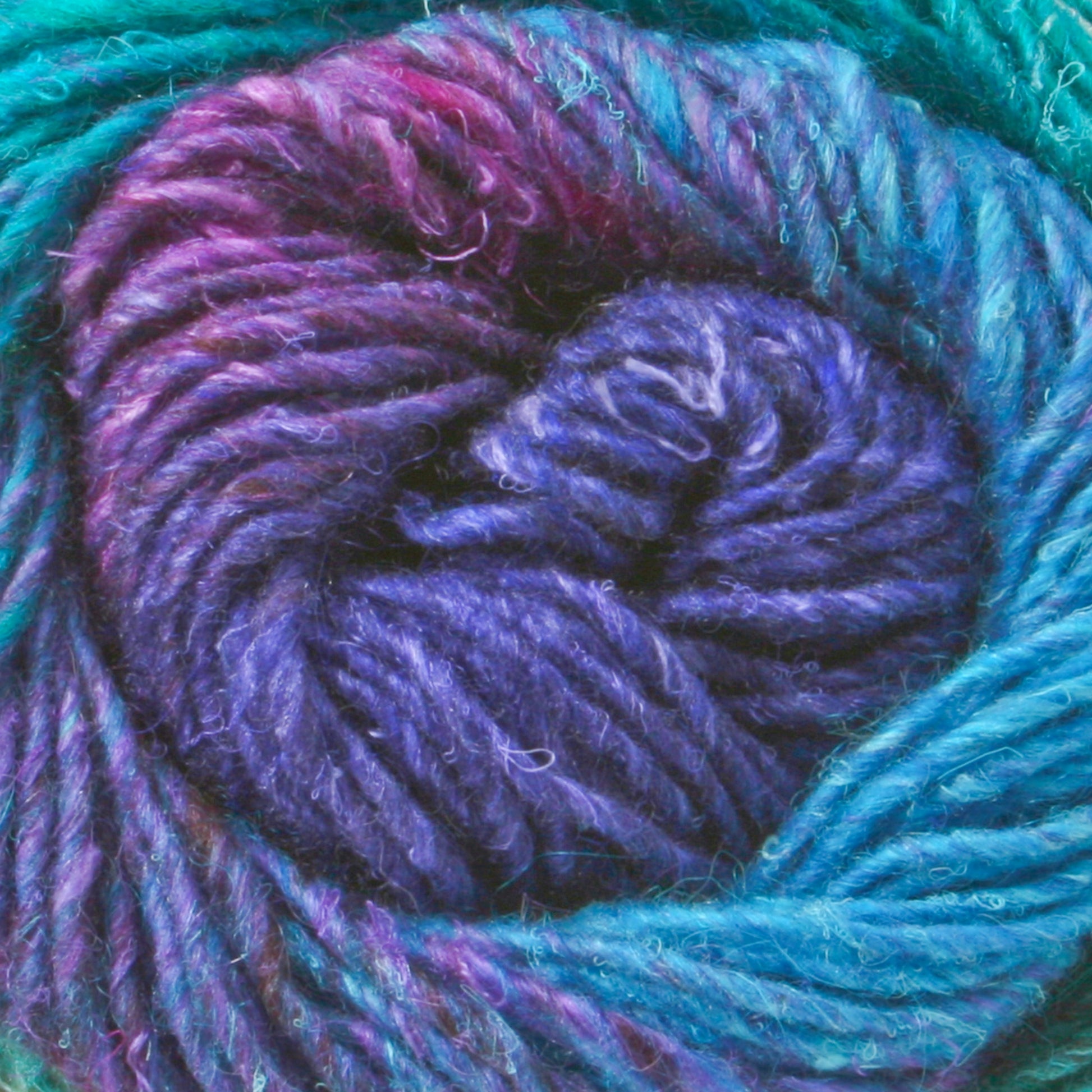 Close-up of a ball of Noro Silk Garden yarn from Knitting Fever/Euro Yarns featuring rich, intertwined colors including shades of blue, purple, and hints of pink and teal. This multi-color self-striping yarn's texture appears soft and slightly fuzzy.